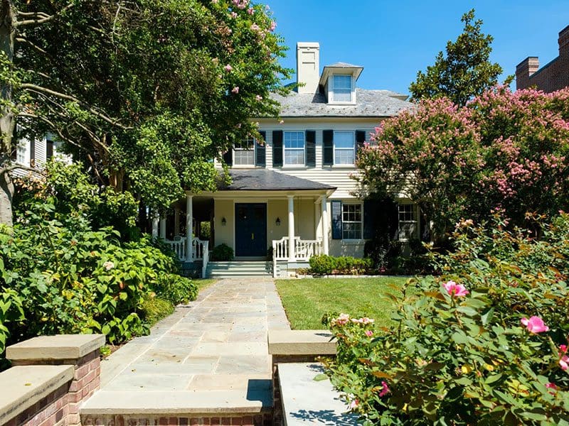 A two-story traditional house with white siding, ideal for real estate buyers, surrounded by lush greenery and blooming pink shrubs, featuring a stone pathway leading to a covered porch.