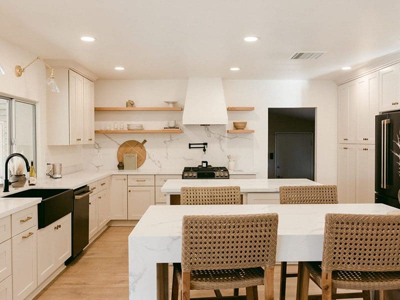 Modern kitchen with white cabinetry, marble countertops, and natural wood accents, designed to appeal to real estate buyers. Includes an island with woven chairs.