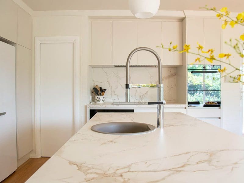 Bright modern kitchen with white marble countertops, white cabinets, a large sink, and yellow flowers in the foreground, perfect for real estate sellers looking to attract buyers.