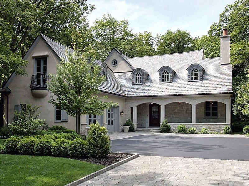 A luxurious two-story house with stone and stucco exterior, ideal for real estate buyers, featuring a paved driveway and surrounded by lush greenery.