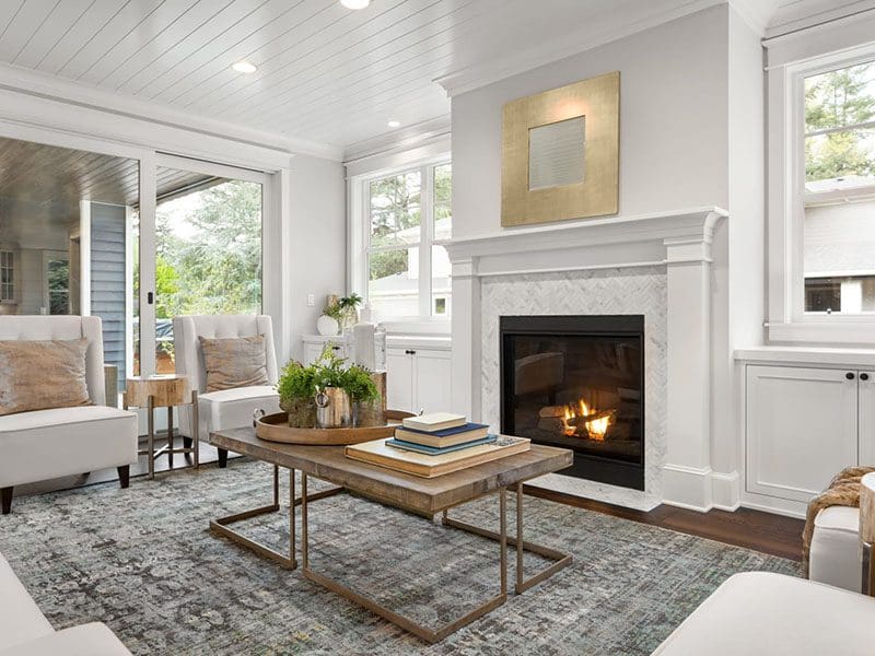 Elegant living room designed for real estate sellers, featuring a lit fireplace, white sofas, wooden coffee table, and a decorative rug, viewed from an angle showcasing bright windows.
