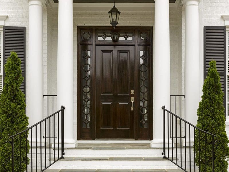 Elegant entrance with a dark wooden door flanked by two white columns, white shutters, and symmetrical topiary shrubs appealing to real estate buyers.