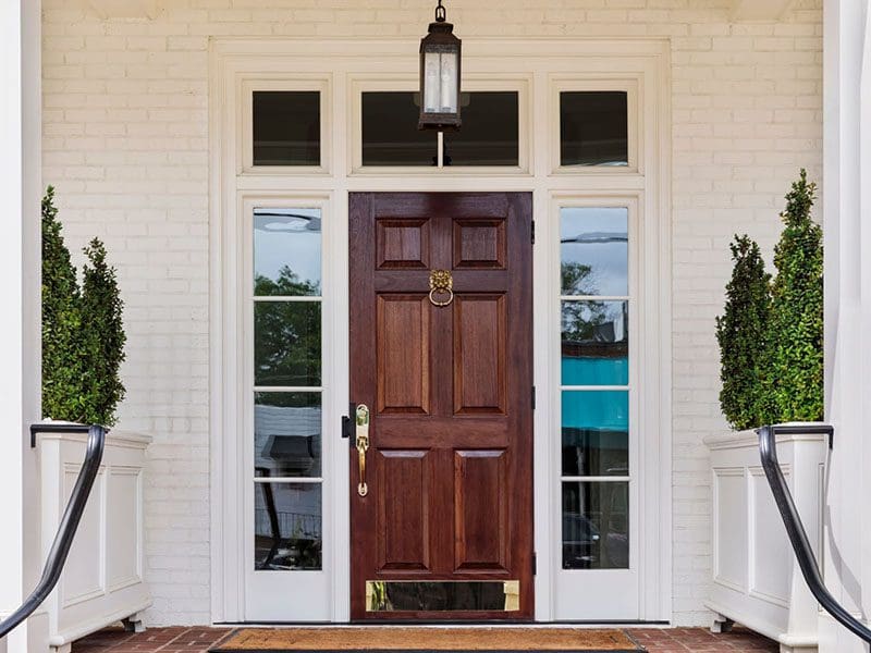 Front view of a wooden door with brass fittings between two potted topiary plants on a porch with a white brick backdrop, appealing to discerning real estate sellers.