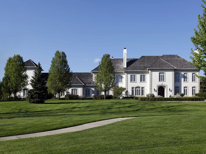 Large white mansion with a gray roof surrounded by manicured green lawns and several trees under a clear blue sky, perfect for discerning real estate buyers.