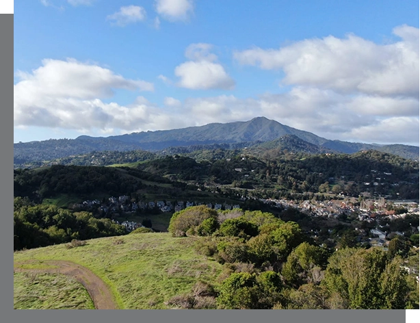 Aerial view of a lush, hilly landscape with a prominent mountain in the background and Corte Madera Real Estate nestled in the valley under a partly cloudy sky.