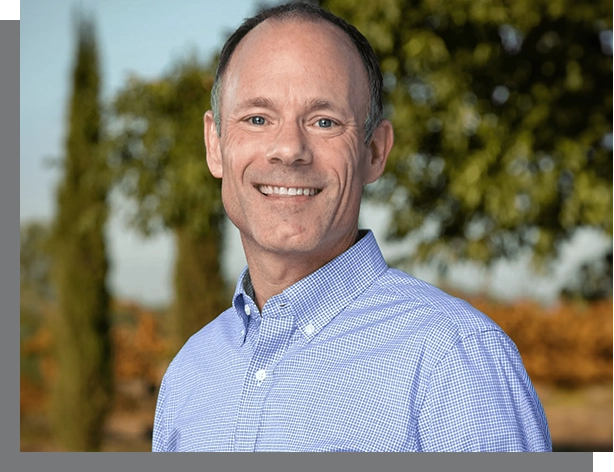 Middle-aged man in a blue checkered shirt smiling outdoors with trees and a clear sky in the background, identified as a real estate coach.