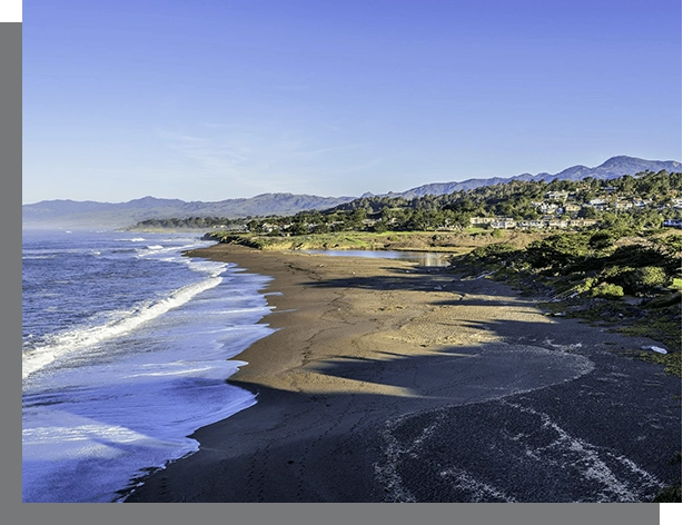 A coastal landscape in Santa Rosa, showing a sandy beach with waves gently touching the shoreline, backed by a green mountain and a clear sky.