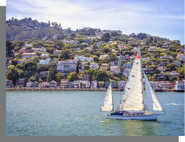 A sailboat glides on the water in front of a hillside dotted with colorful houses under a clear blue sky, showcasing the scenic views typical of San Rafael Real Estate.