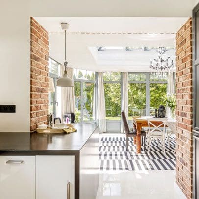 A bright, modern kitchen with exposed brick walls, checkered floor, and a dining area near glass doors leading to a garden, perfect for real estate buyers.
