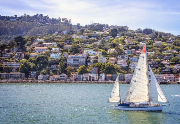 A sailboat with white sails glides on water in front of a hillside town with densely packed colorful houses under a clear sky, ideal for those engaged in real estate property search.
