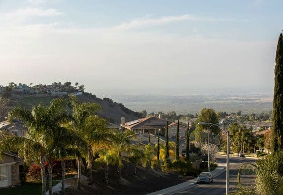A scenic overlook of a suburban neighborhood featuring real estate properties, palm trees, houses, and hills under a clear sky at sunset.